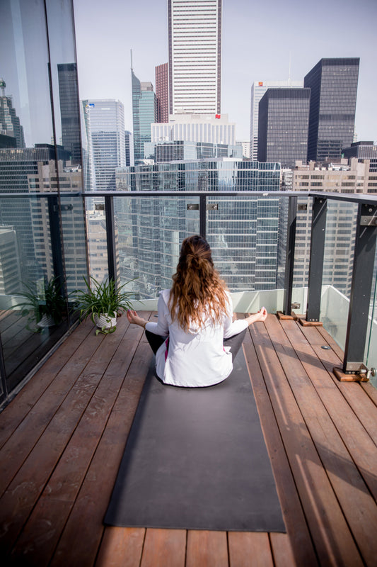Person sits facing away from the camera on a yoga mat that is on a balcony with a wooden floor. You can see the city in the distance.